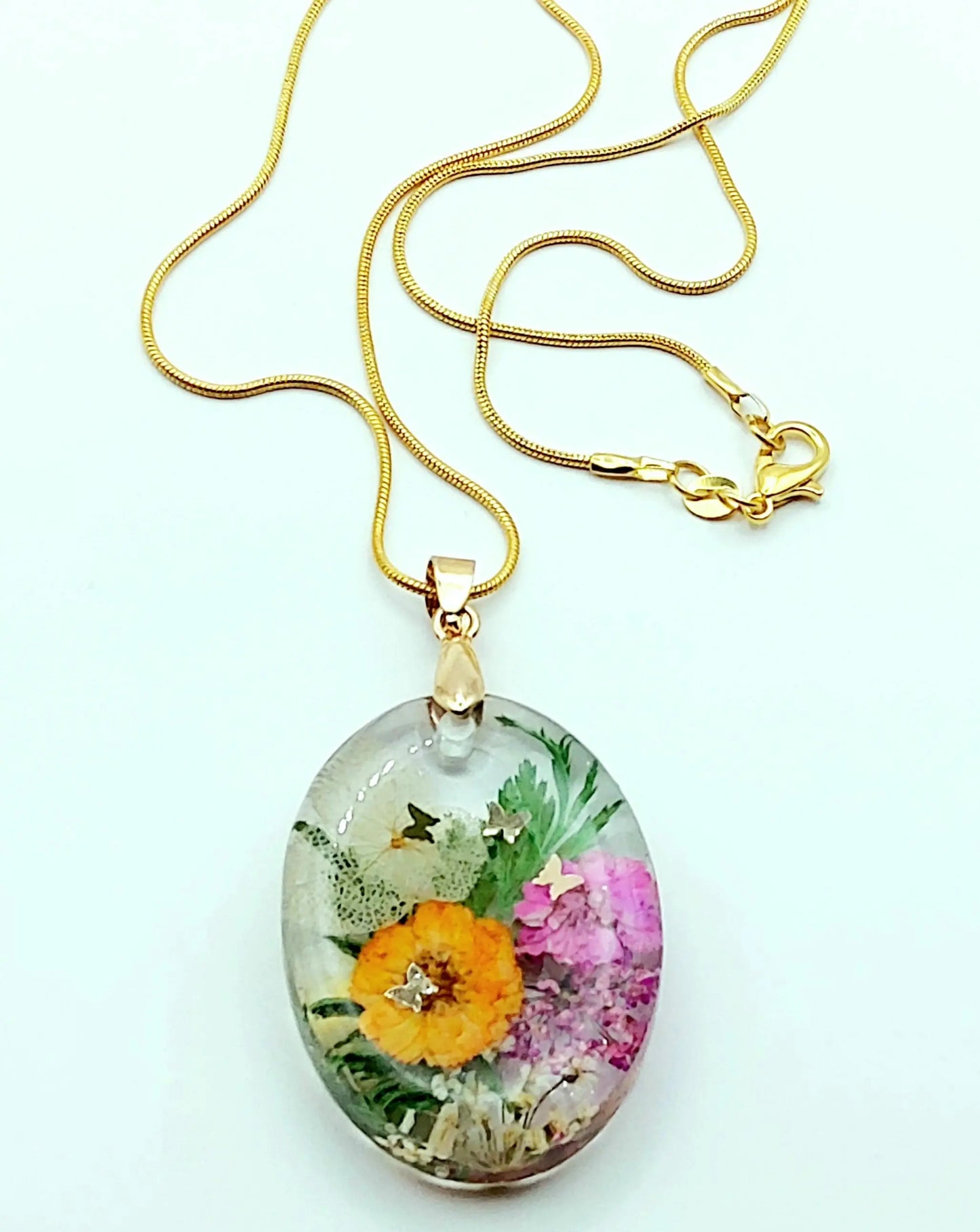 Pendentif Ovale en résine époxy et sa chaîne dorée. Fleurs multicolores et papillons dorés. Plan large.