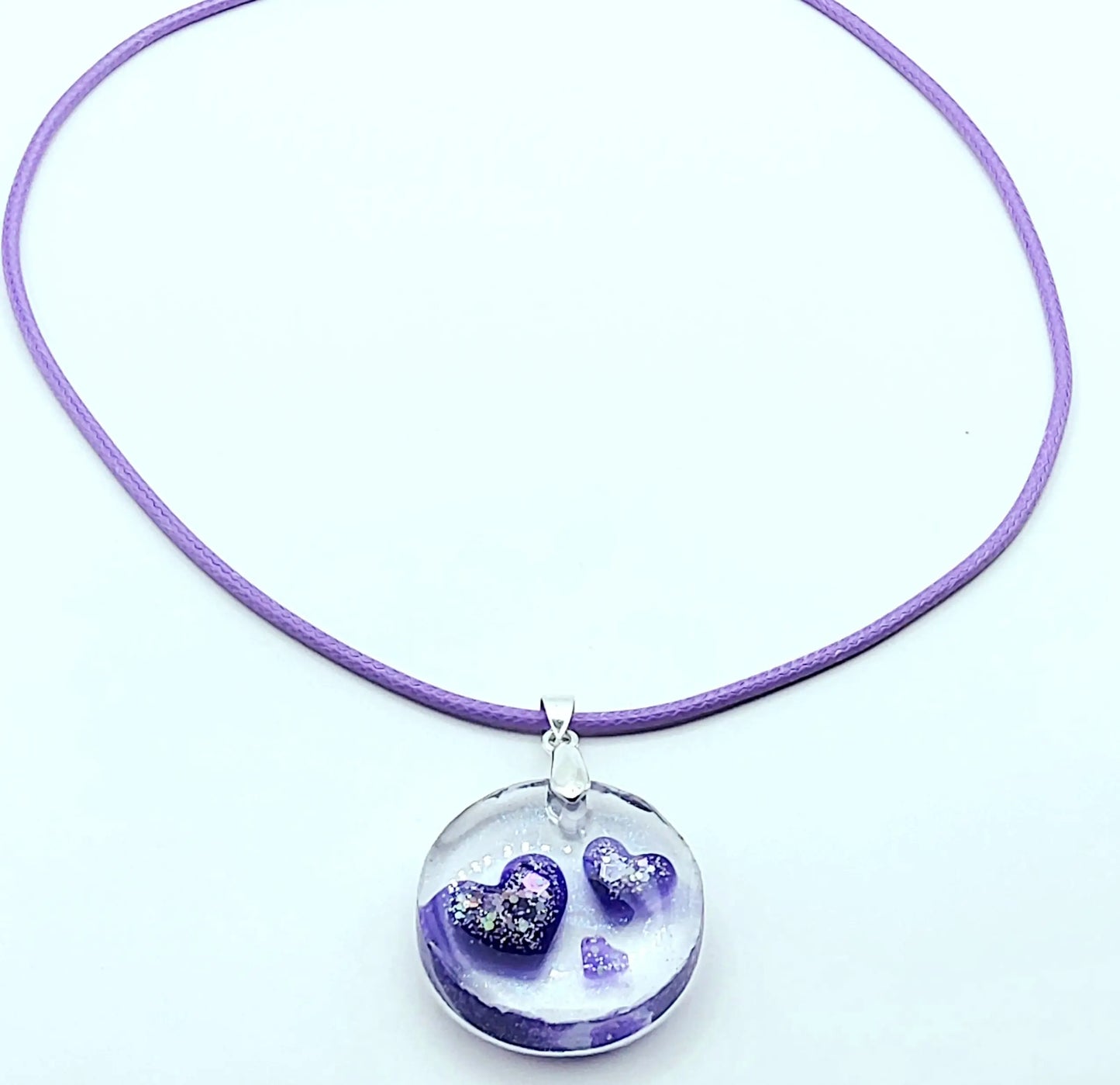 Pendentif ROND en résine époxy. Transparent avec effet irisé. Renferme 3 petits cœurs violets et paillettes argentées. Cordon en coton ciré mauve. PRETTY RESINE