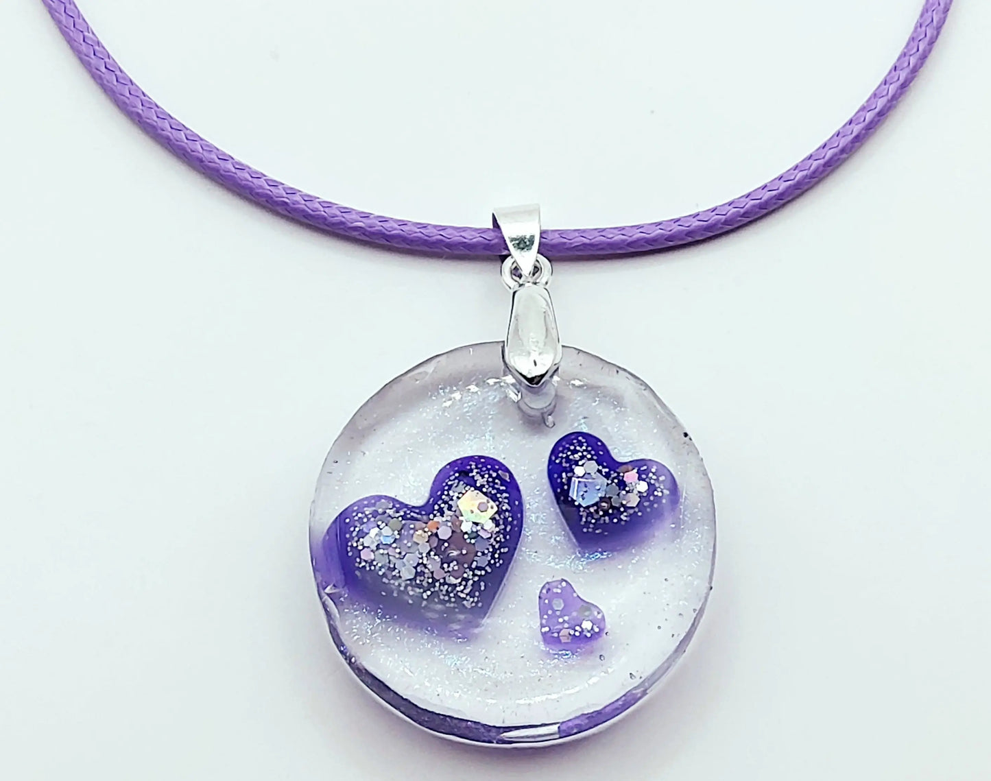 Pendentif ROND en résine époxy. Transparent avec effet irisé. Renferme 3 petits cœurs violets et paillettes argentées. Cordon en coton ciré mauve. PRETTY RESINE