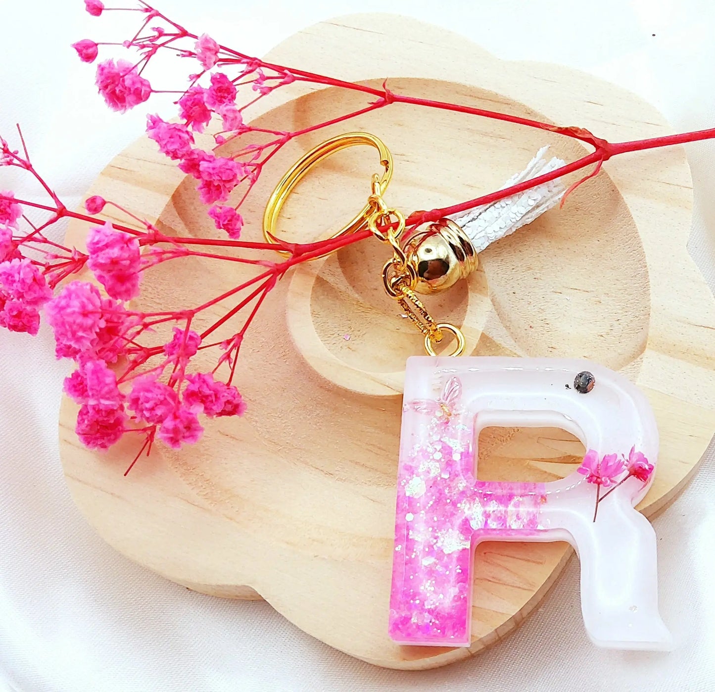Porte-clés LETTRE R en résine époxy. Fond blanc. Paillettes, fleurs et papillon rose. Pompon gland pailleté blanc. PRETTY RESINE