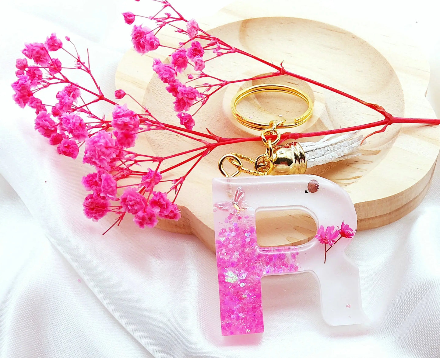 Porte-clés LETTRE R en résine époxy. Fond blanc. Paillettes, fleurs et papillon rose. Pompon gland pailleté blanc. PRETTY RESINE