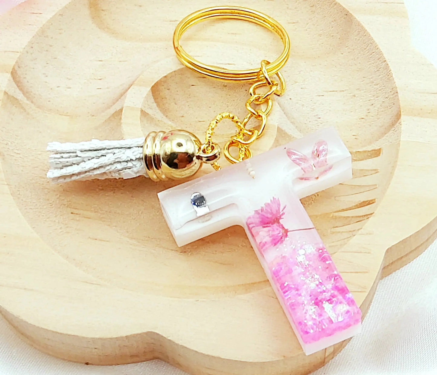 Porte-clés LETTRE T en résine époxy. Fond blanc, paillettes, fleurs et papillon roses. Pompon gland pailleté blanc. PRETTY RESINE
