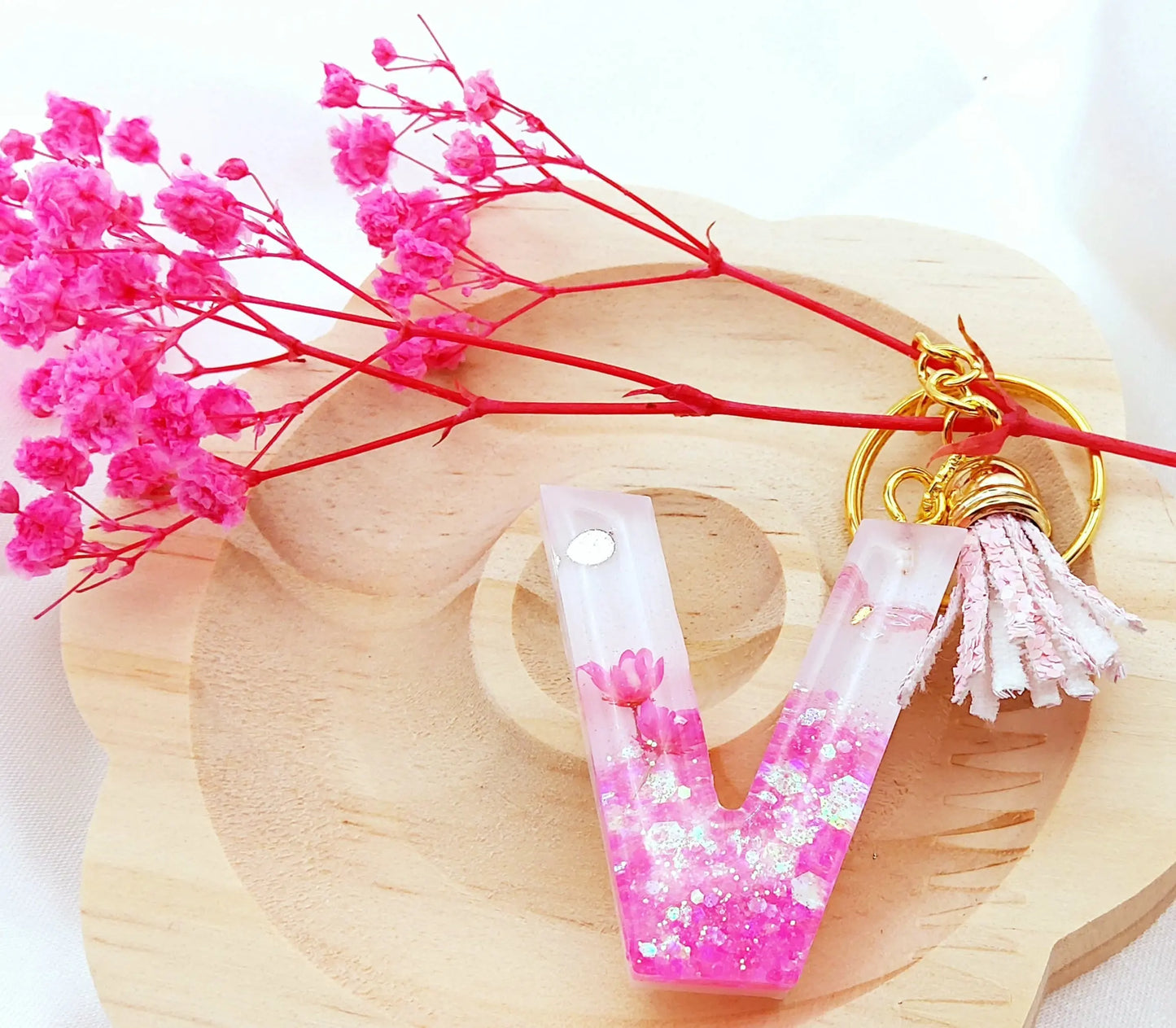 Porte-clés LETTRE V en résine époxy. Fond blanc. Paillettes, fleurs et papillon roses. Pompon gland pailleté rose. PRETTY RESINE