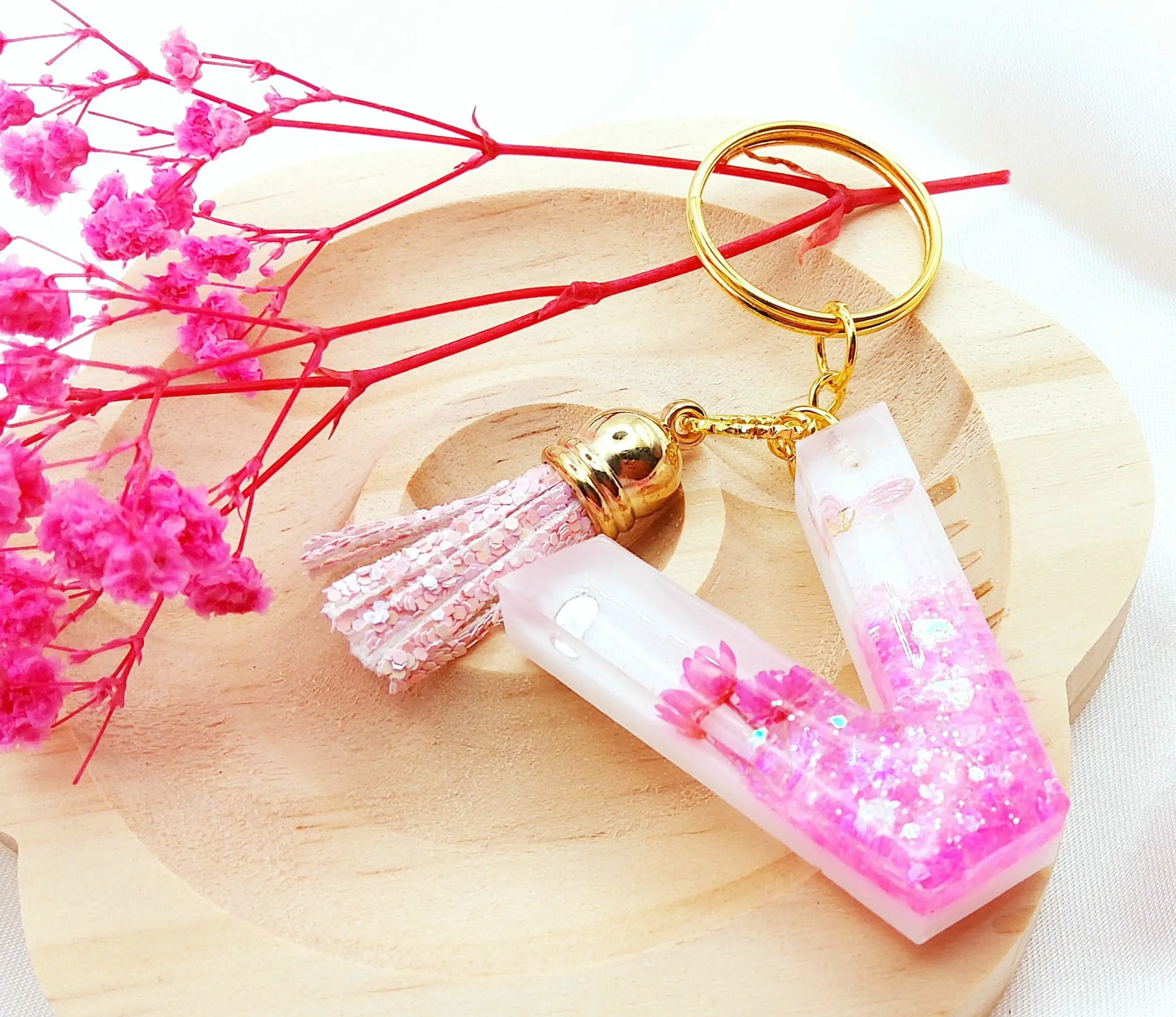Porte-clés LETTRE V en résine époxy. Fond blanc. Paillettes, fleurs et papillon roses. Pompon gland pailleté rose. PRETTY RESINE