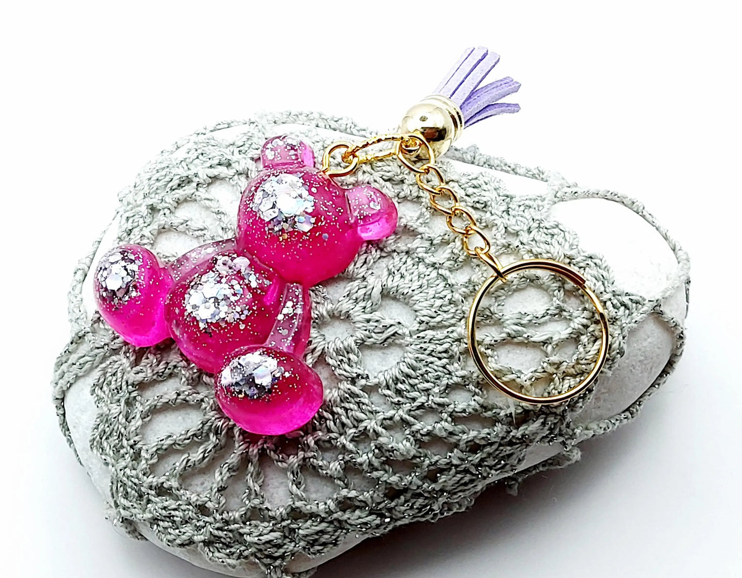 Porte-clés "Ourson assis" en résine époxy, rose et paillettes argentées. Anneau doré, pompon gland mauve. Posé sur un galet.