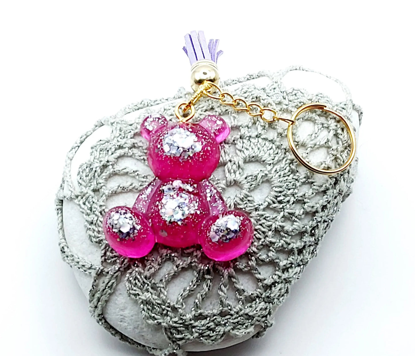 Porte-clés "Ourson assis" en résine époxy, rose et paillettes argentées. Anneau doré, pompon gland mauve. Posé sur un galet.