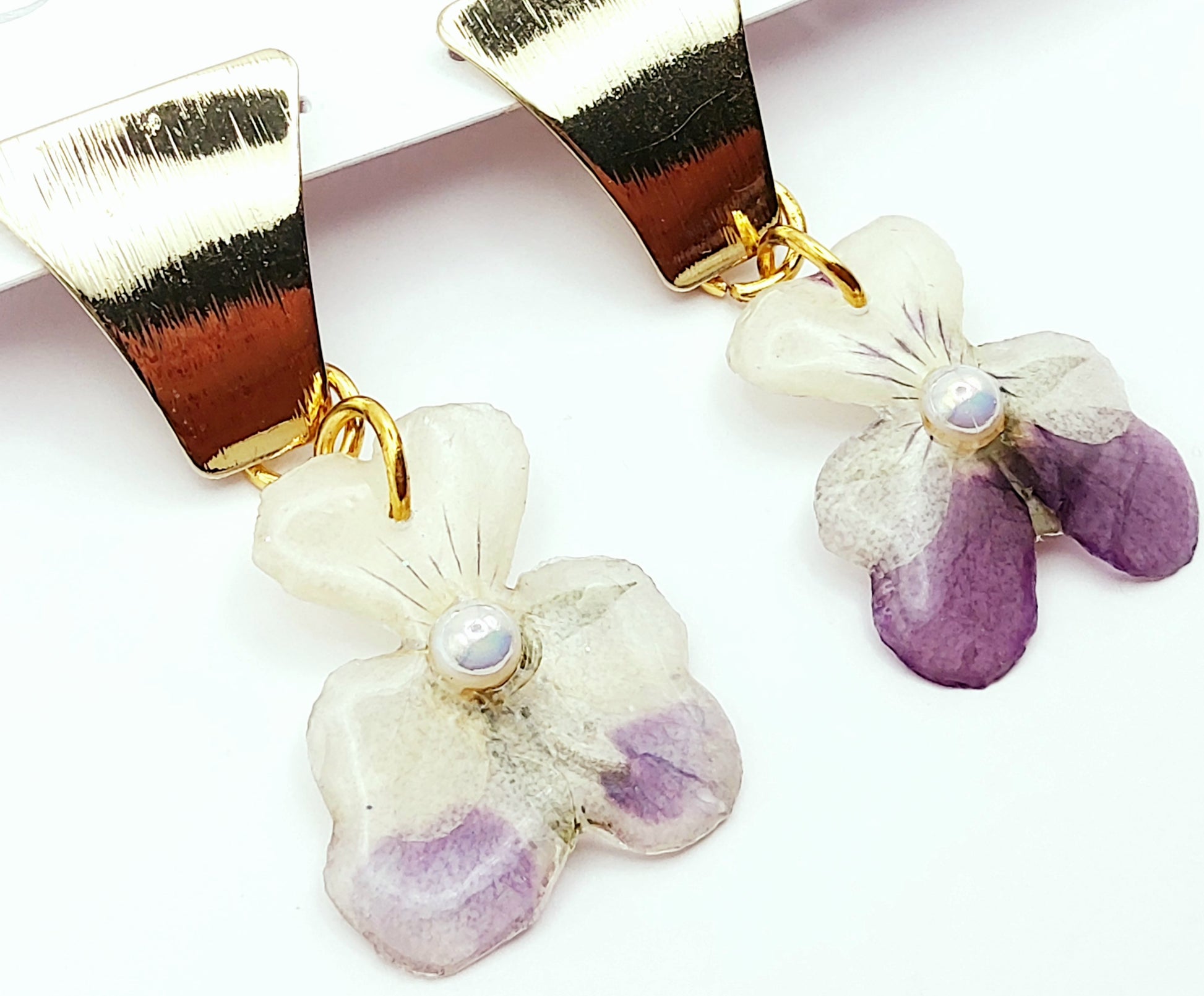 Boucles d'oreilles Fleurs de pensées en résine. Violet et blanc avec une perle blanche au centre. Apprêt doré.