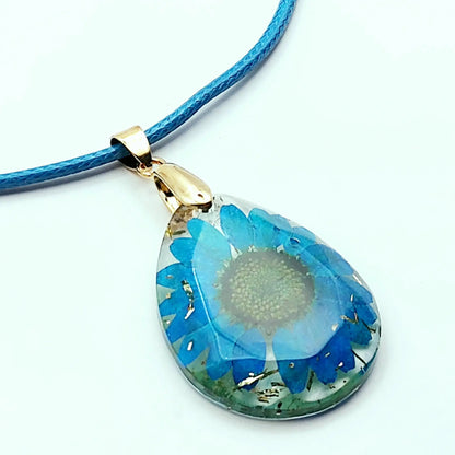 Pendentif OVALE en résine époxy. Fleur bleue et paillettes dorées. Cordon en coton ciré bleu.