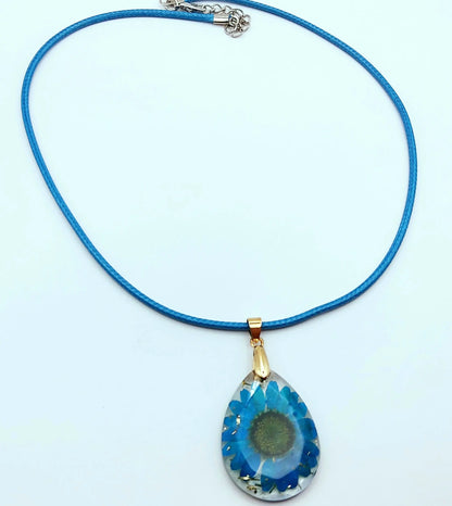 Pendentif OVALE en résine époxy. Fleur bleue et paillettes dorées. Cordon en coton ciré bleu.