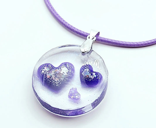 Pendentif ROND en résine époxy. Transparent avec effet irisé. Renferme 3 petits cœurs violets et paillettes argentées. Cordon en coton ciré mauve. PRETTY RESINE