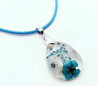 Pendentif "Goutte d'eau" en résine époxy,. Transparent avec fleurs bleues. Cordon en coton ciré bleu clair. Gros plan.