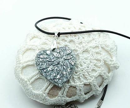 Pendentif "Petit Coeur" en résine époxy, paillettes argentées, cordon en coton ciré noir. Posé sur un galet.