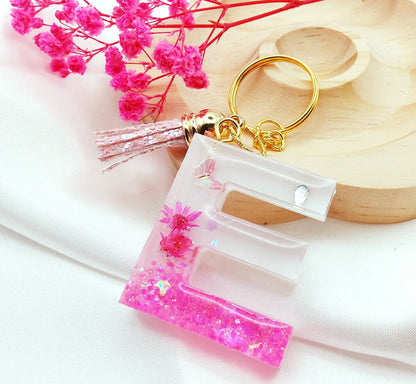 Porte-clés LETTRE E en résine époxy. Fond blanc. Paillettes, fleurs et papillon roses. Pompon gland pailleté rose. PRETTY RESINE