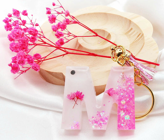 Porte-clés LETTRE M en résine époxy. Fond blanc. Paillettes, fleurs et papillon roses. Pompon gland rose. PRETTY RESINE