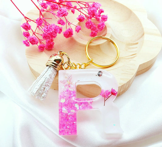 Porte-clés LETTRE R en résine époxy. Fond blanc. Paillettes, fleurs et papillon rose. Pompon gland pailleté blanc. PRETTY RESINE