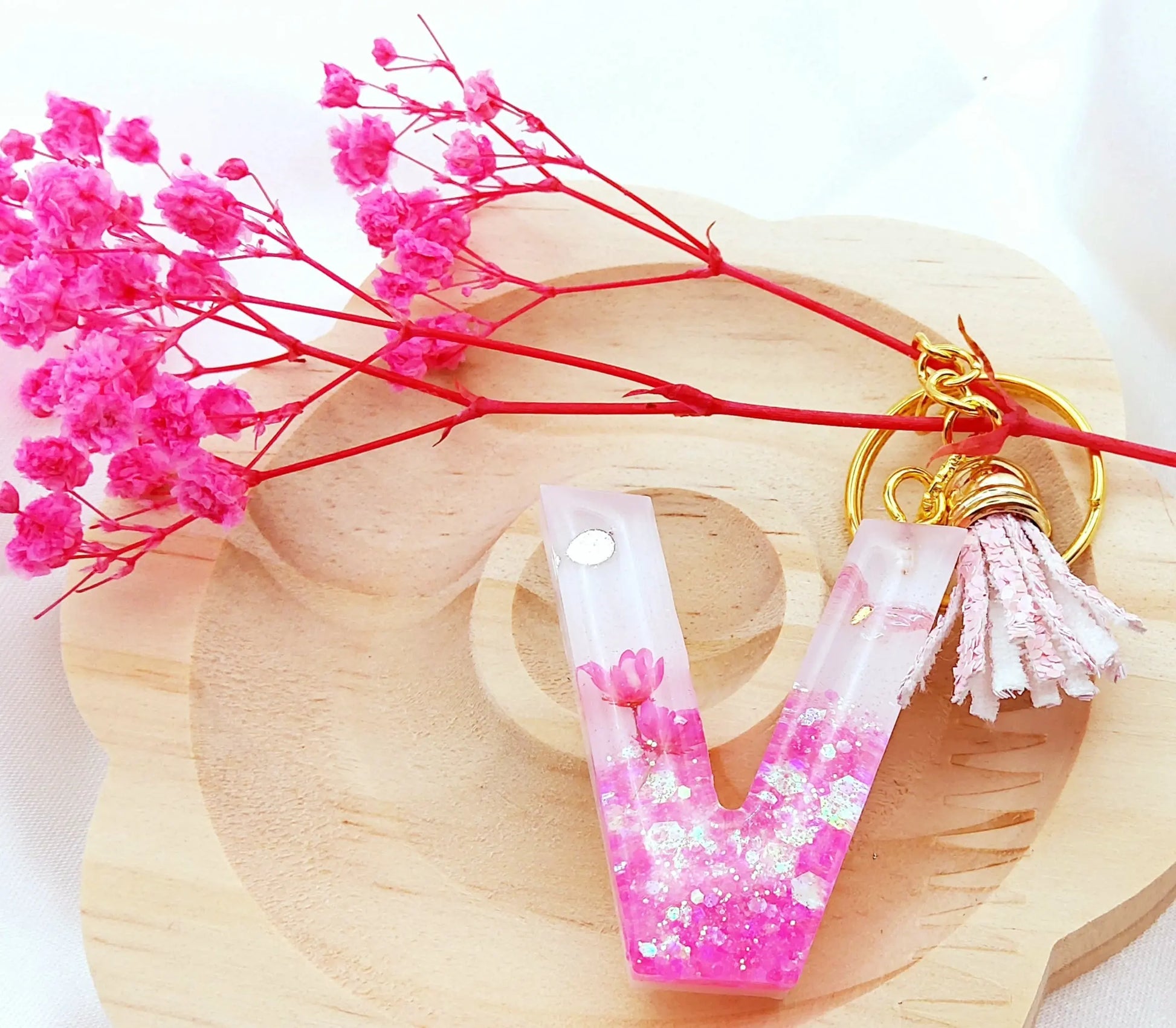 Porte-clés LETTRE V en résine époxy. Fond blanc. Paillettes, fleurs et papillon roses. Pompon gland pailleté rose. PRETTY RESINE