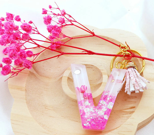 Porte-clés LETTRE V en résine époxy. Fond blanc. Paillettes, fleurs et papillon roses. Pompon gland pailleté rose. PRETTY RESINE