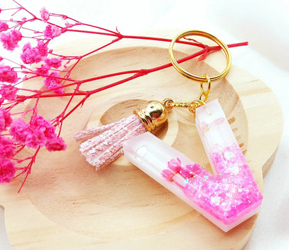 Porte-clés LETTRE V en résine époxy. Fond blanc. Paillettes, fleurs et papillon roses. Pompon gland pailleté rose. PRETTY RESINE