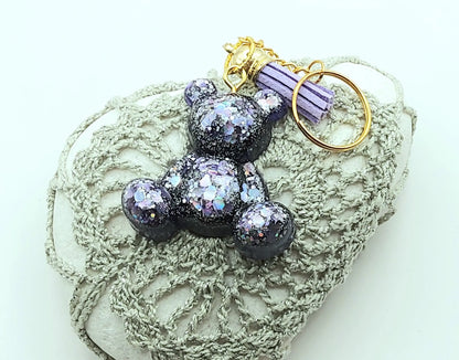 Porte-clés "Ourson assis" en résine époxy, violet et paillettes mauves. Anneau doré, pompon gland mauve. Posé sur un galet.