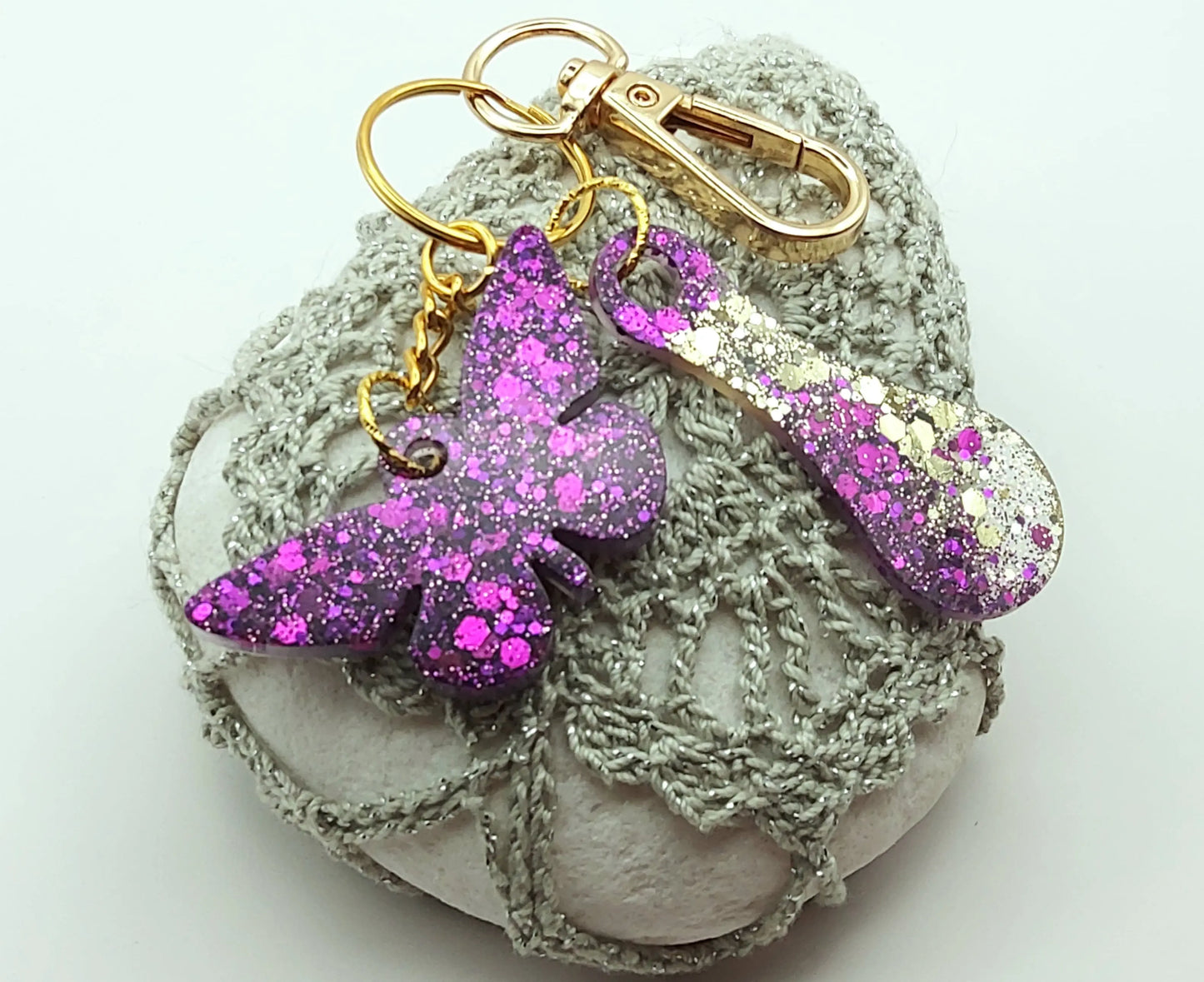 Porte-clés "Papillon et jeton de caddie" en résine époxy, paillettes violettes, anneau et mousqueton dorés, posé sur un galet