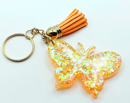 Porte-clés "Papillon" en résine époxy. Paillettes oranges et jaunes. Anneau doré. Pompon gland orange.