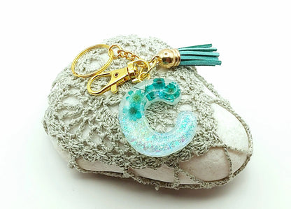 Porte-clés "Petite Lettre C" en résine époxy, fleurs et pompon gland turquoise, posé sur un galet