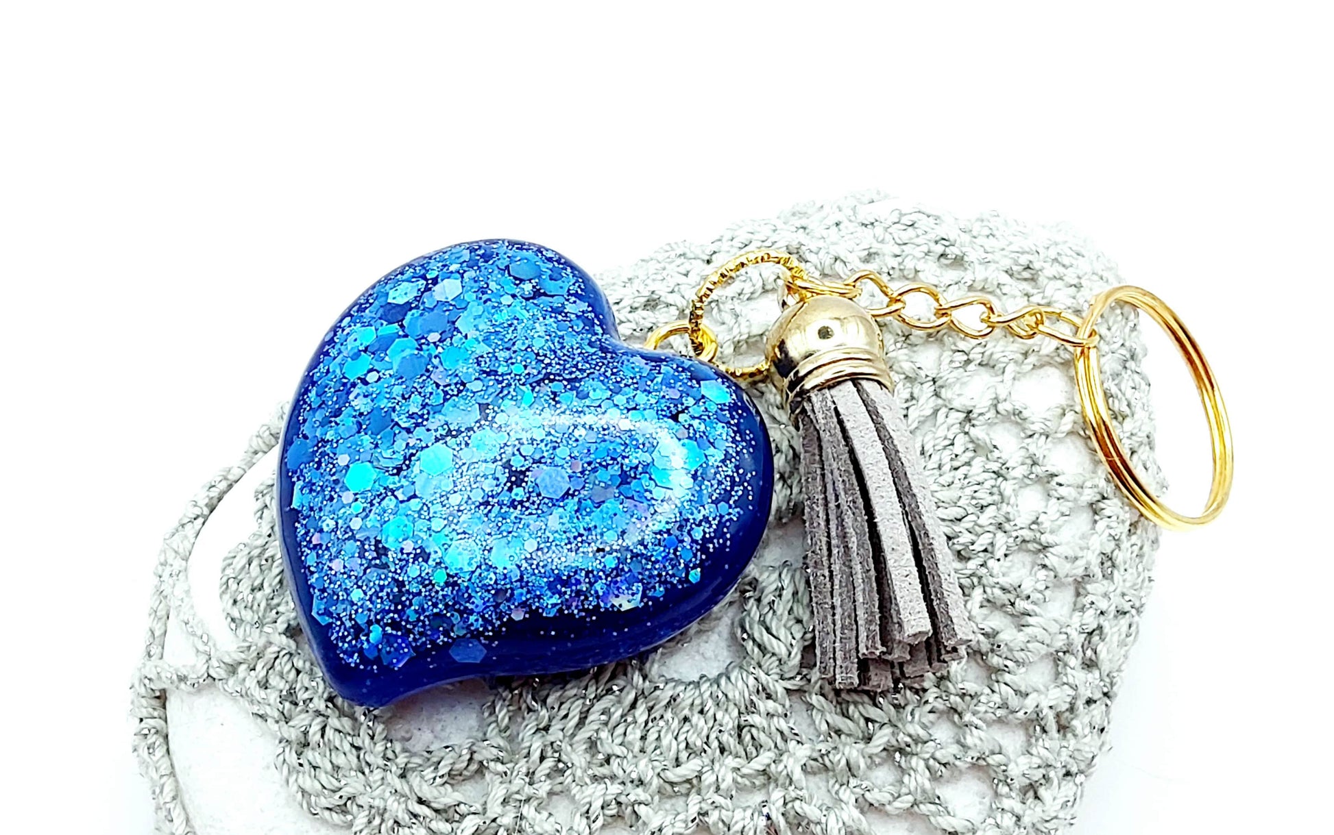 Porte-clés "Gros Coeur" en résine époxy. Paillettes bleues, pompon gland gris clair, anneau doré. Posé sur un galet.