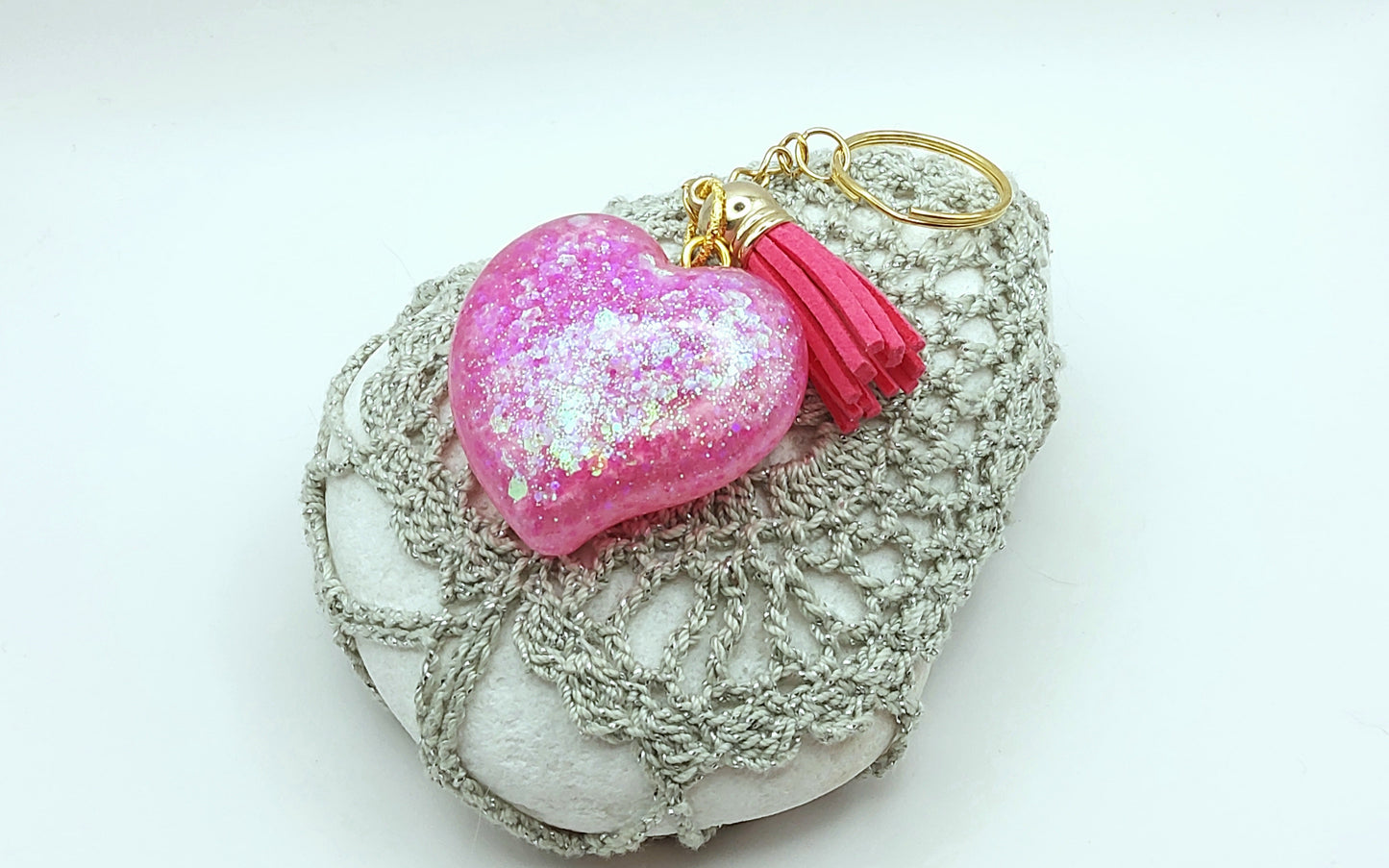 Porte-clés "Gros Coeur" en résine époxy, paillettes holographiques roses, pompon gland rose fuchsia, anneau doré. Posé sur un galet.