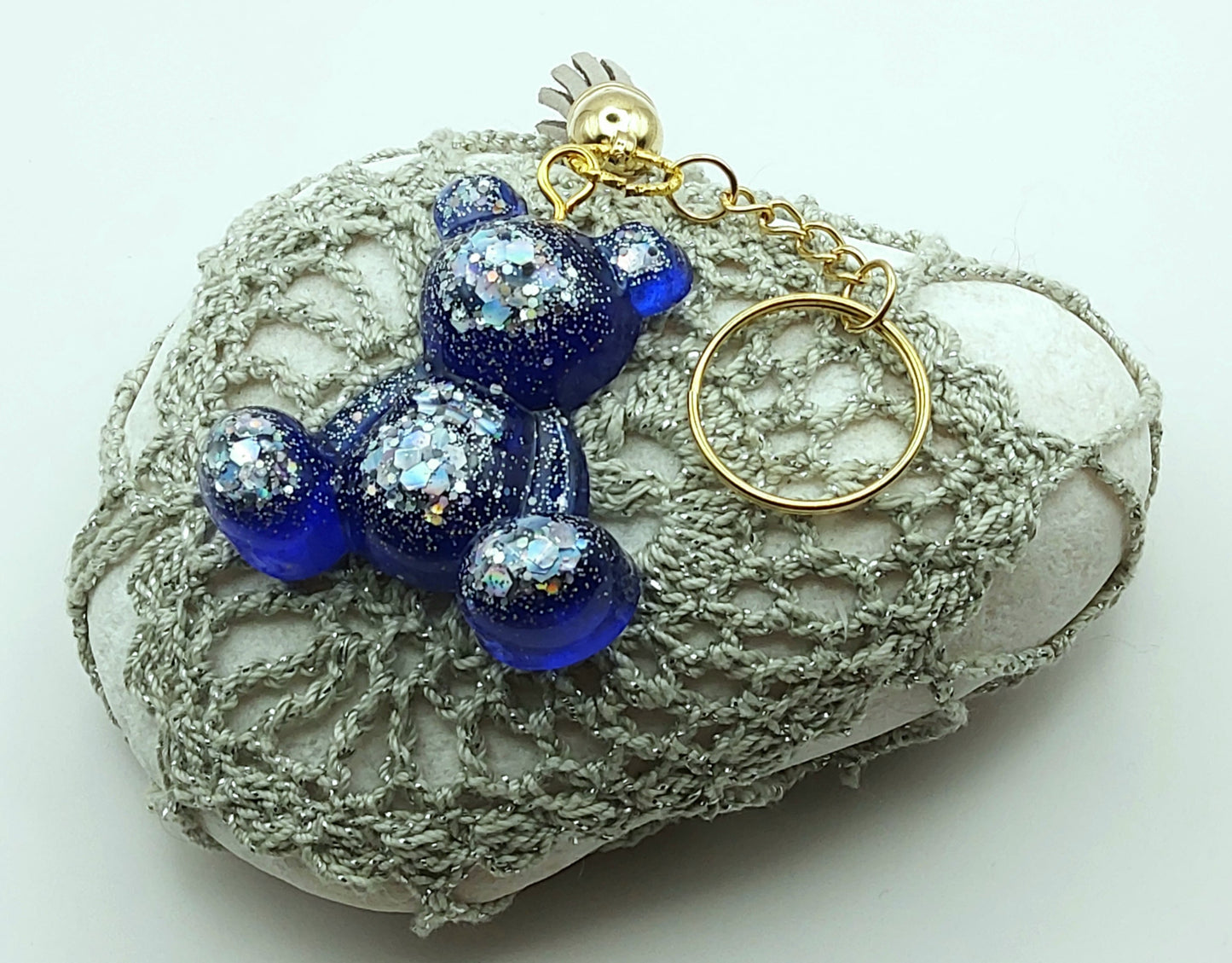 Porte-clés "Ourson assis" en résine époxy, bleu marine et paillettes argentées. Anneau doré. Pompon gland gris clair. Posé sur un galet.