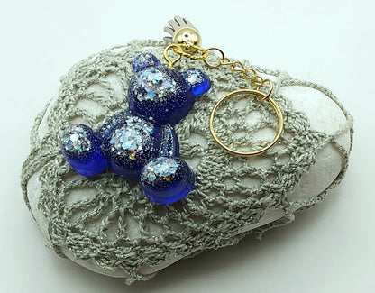 Porte-clés "Ourson assis" en résine époxy, bleu marine et paillettes argentées. Anneau doré. Pompon gland gris clair. Posé sur un galet.