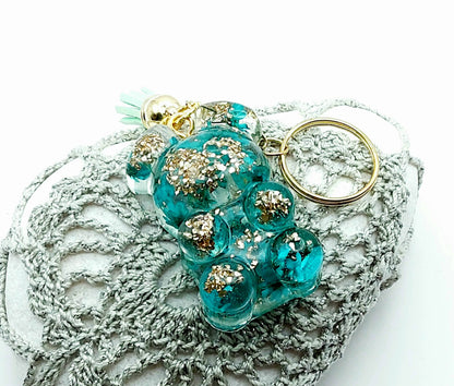 Porte-clés "Ourson debout" en résine époxy. Fleurs turquoises et verre pilé doré. Anneau doré et pompon gland vert pastel. Posé sur un galet.