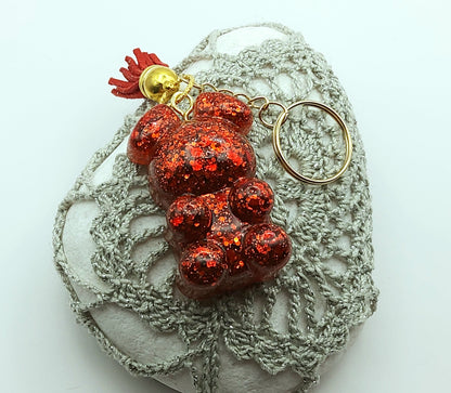 Porte-clés "Ourson debout" en résine époxy, paillettes rouges, pompon gland rouge, anneau doré. Posé sur un galet.