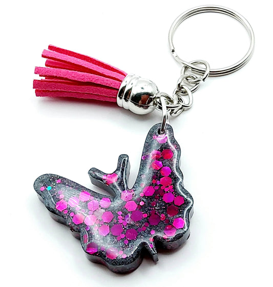 Porte-clés "Papillon" en résine époxy, gris et paillettes fuchsia. Anneau argenté. Pompon gland fuchsia.