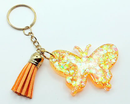 Porte-clés "Papillon" en résine époxy. Paillettes oranges et jaunes. Anneau doré. Pompon gland orange.